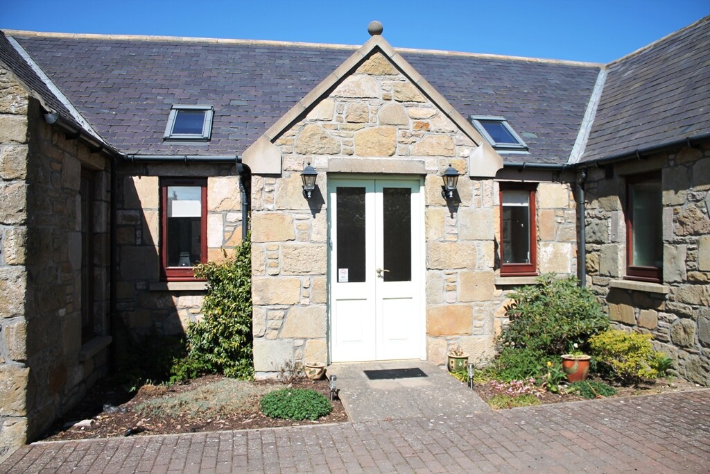 The exterior of the cottage displays a stone facade framed by shrubs and flower beds. A pair of lanterns adorns either side of the entrance, which is a white double door. Above, skylights are integrated into the sloping roof under a clear blue sky.