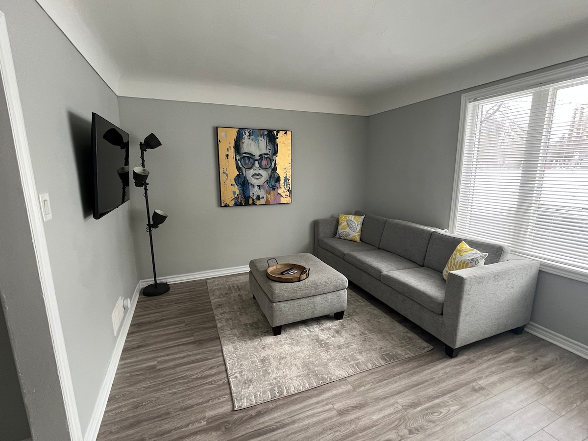 A cozy living area features a light grey sofa accompanied by yellow-accented cushions. A rectangular ottoman rests on a neutral-toned rug. A television is mounted on the wall, and natural light filters in through a window adorned with blinds.