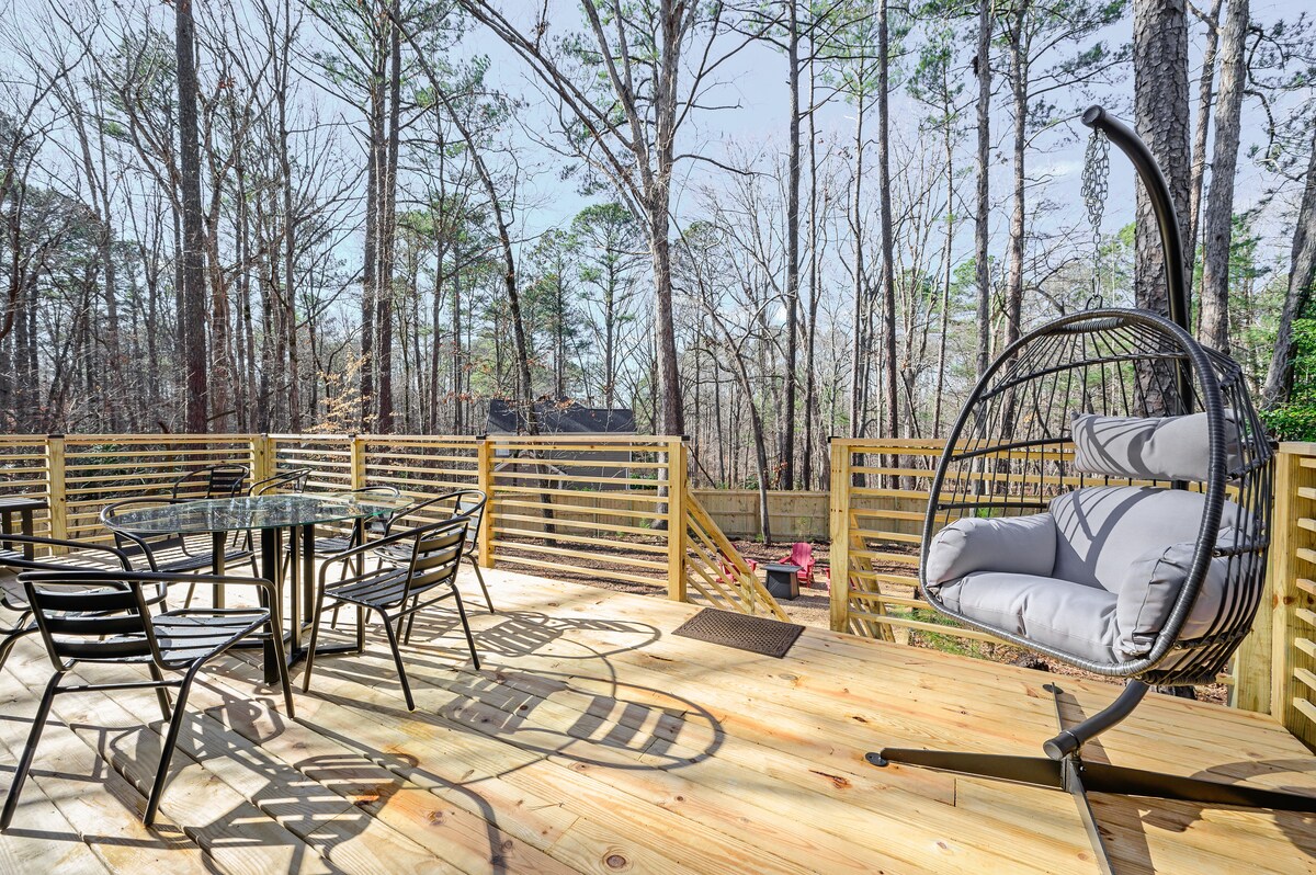 An outdoor dining area features a round table surrounded by six slender black chairs. A large hanging chair sits nearby, offering a cushioned spot for relaxation. The deck is surrounded by wooden railings and overlooks a serene wooded landscape with tall trees.