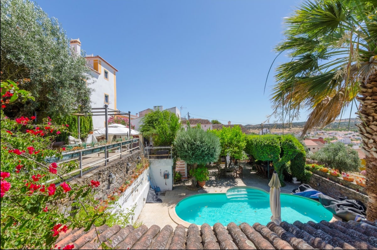 The outdoor area features a private pool surrounded by lush greenery. A sun umbrella stands near the pool, with vibrant flowers adding color. The hilltop setting provides panoramic views of the countryside, while a charming house is visible in the background.
