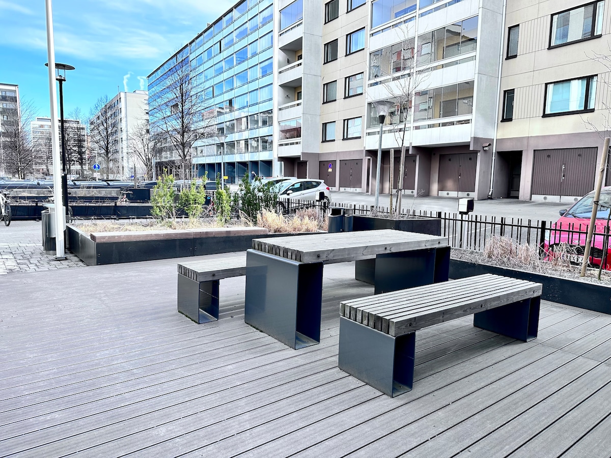 An outdoor seating area is presented, featuring a rectangular table made of wood and metal, accompanied by two benches. The space is surrounded by planters with low shrubs, and the background showcases modern apartment buildings along a sidewalk.