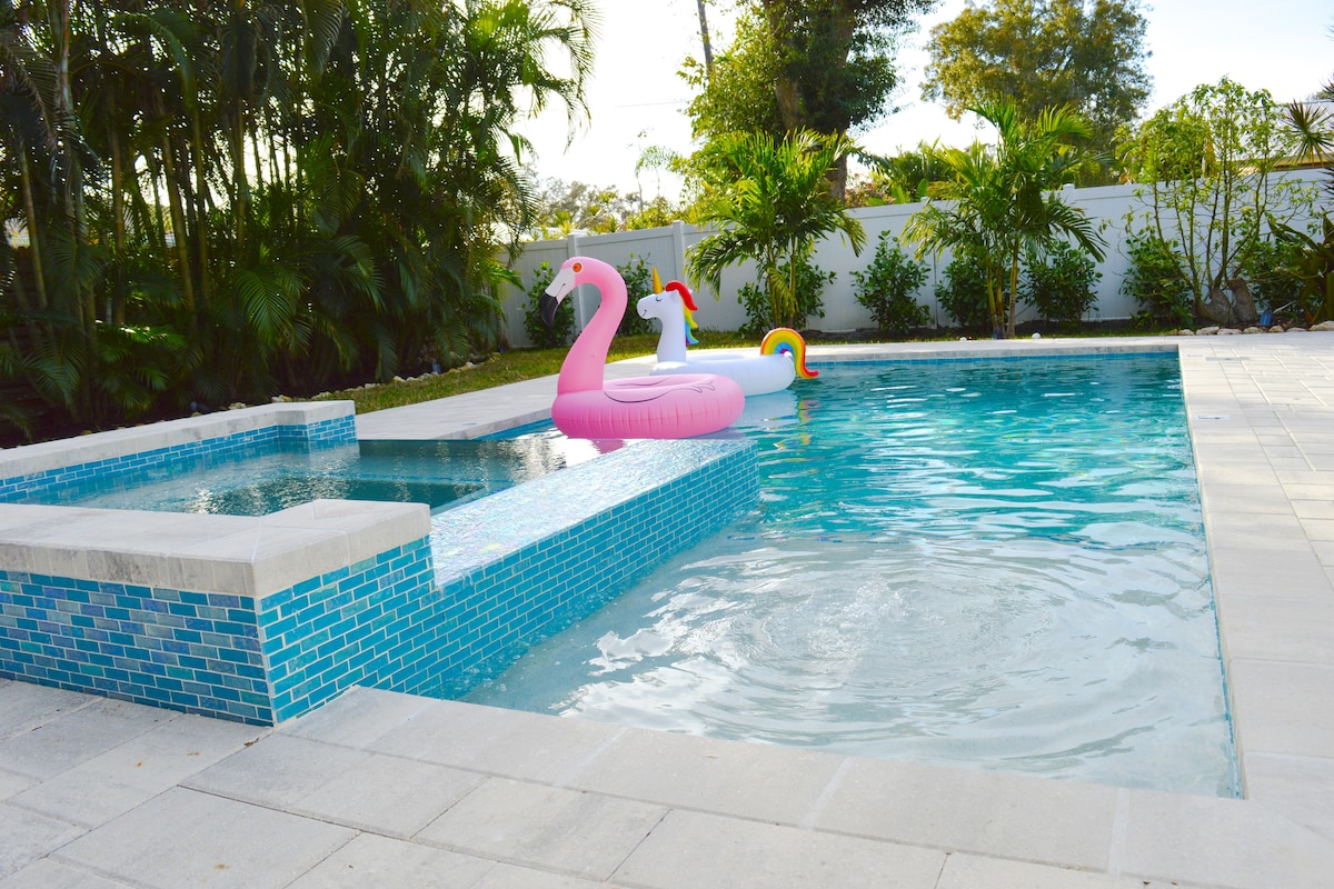 A serene saltwater pool features a spa section, adorned with colorful inflatable floats. Lush greenery surrounds the pool area, enhancing the tranquil outdoor space, with a clean, modern stone deck complementing the vibrant tile accents.