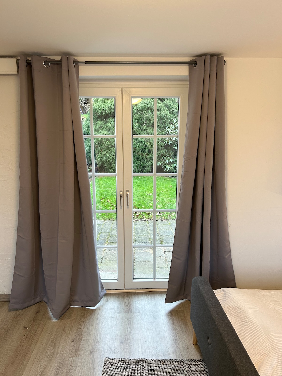 A set of double windows with clear glass panes is framed by light gray curtains, allowing natural light to enter the room. Beyond the windows, a green lawn and mature shrubs are visible, complementing the serene environment.