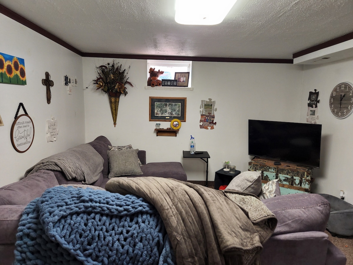 A welcoming living space features a large sectional sofa adorned with decorative pillows and throws. A television is positioned against the wall, and various artwork and decor items are placed around the room, creating an inviting atmosphere. A small window allows light to enter.
