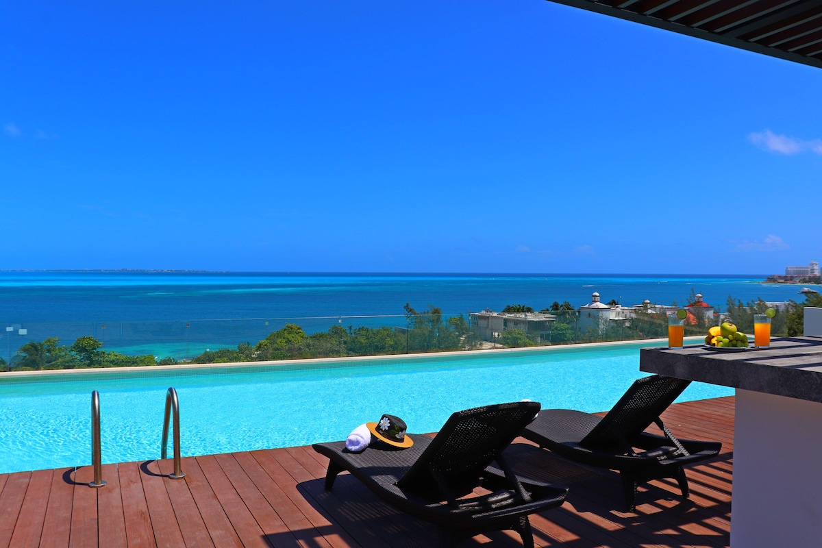 An infinity pool stretches across the image, offering stunning views of the turquoise waters of the Caribbean Sea. Two lounge chairs are positioned nearby, with refreshments set on a table. The clear blue sky and serene landscape enhance the tranquil atmosphere.