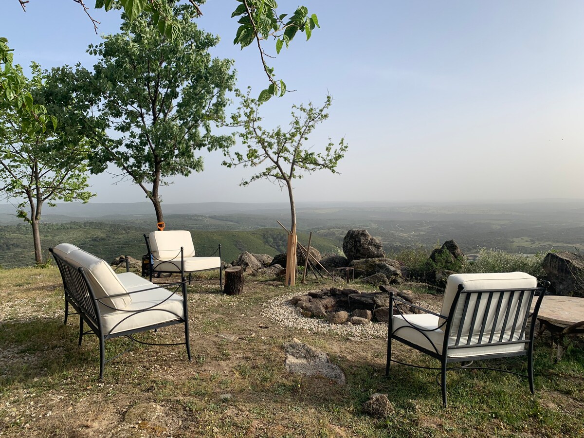 A seating area features light-colored chairs arranged around a fire pit, with a view of rolling hills and trees in the background. The landscape is open and expansive, illustrating the tranquility of the natural surroundings.