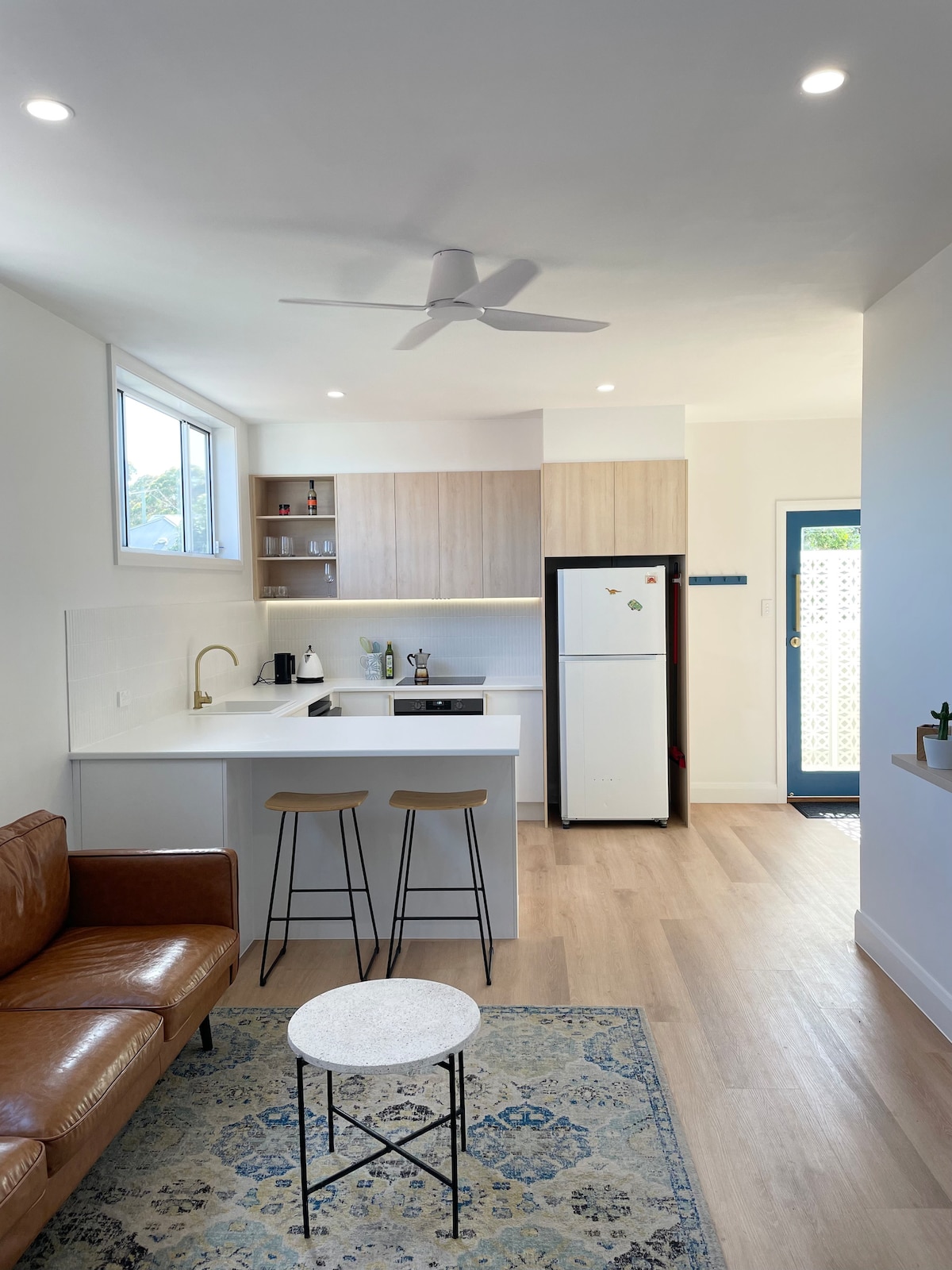A modern kitchen space is shown, featuring light wood cabinetry and a white countertop. Two stools are positioned at the kitchen island, with a small round table nearby. A comfortable brown leather couch and a door that leads outside can be seen, enhancing the open layout.