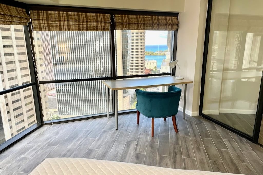Large floor-to-ceiling windows offer a view of the ocean and cityscape. A wooden desk complements a green accent chair in the corner. Natural light fills the spacious room, highlighting the modern tile flooring and neutral-toned decor.