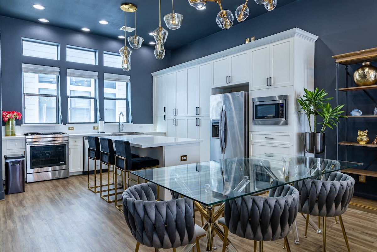 A contemporary kitchen and dining area features sleek white cabinetry and modern stainless steel appliances. A glass dining table is surrounded by upholstered chairs with a tufted design. Pendant lights hang from the ceiling, illuminating the space with a soft glow.