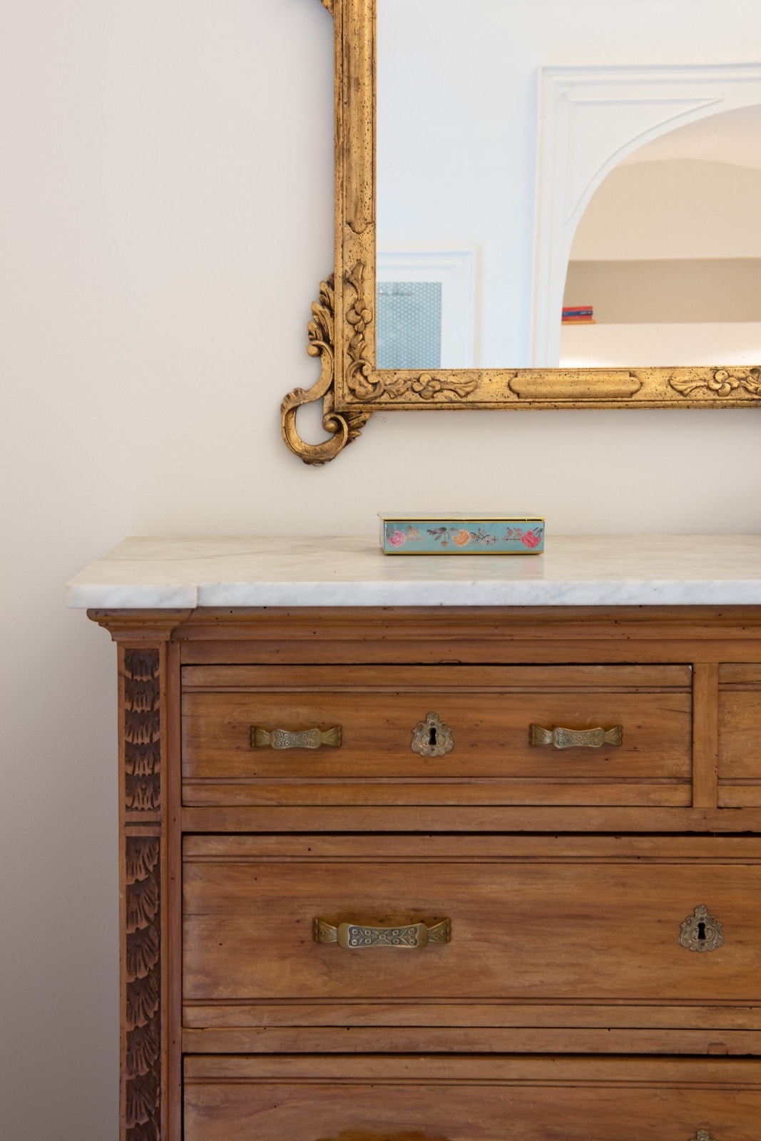 A wooden dresser with intricate carvings is displayed beneath an ornately framed mirror. The dresser features several drawers with brass handles, and a decorative box rests on the marble countertop, reflecting a sense of understated elegance.