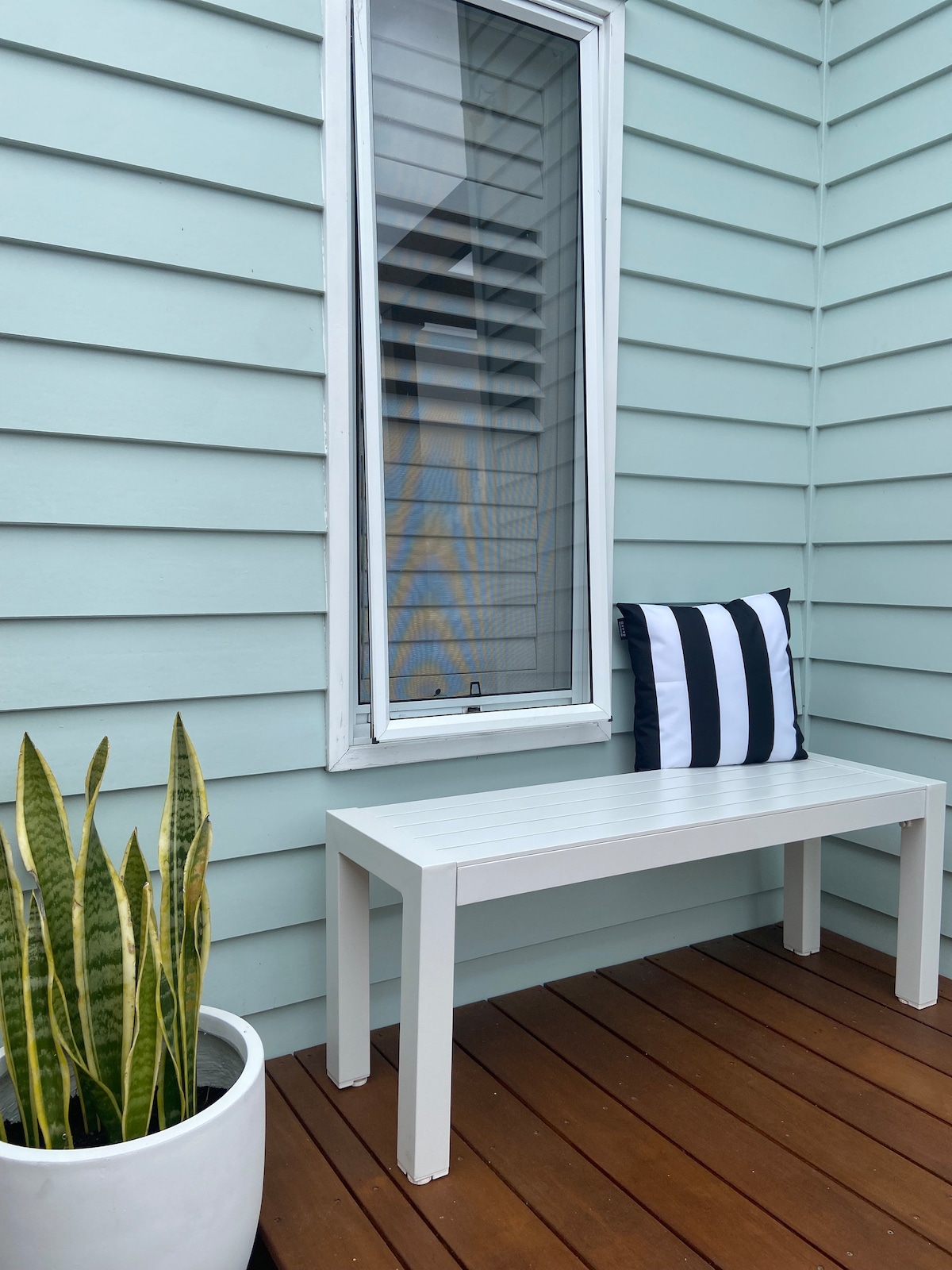 A small decked area features a white bench against a light blue wall, with a striped black and white cushion adding a touch of comfort. A potted snake plant stands nearby, complementing the serene setting.