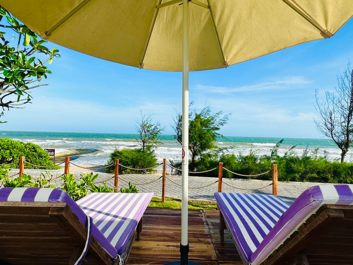 Two lounge chairs with striped cushions face the ocean, placed beneath a large cream umbrella. The view features gentle waves and a cloudy sky, with greenery in the foreground enhancing the coastal setting.