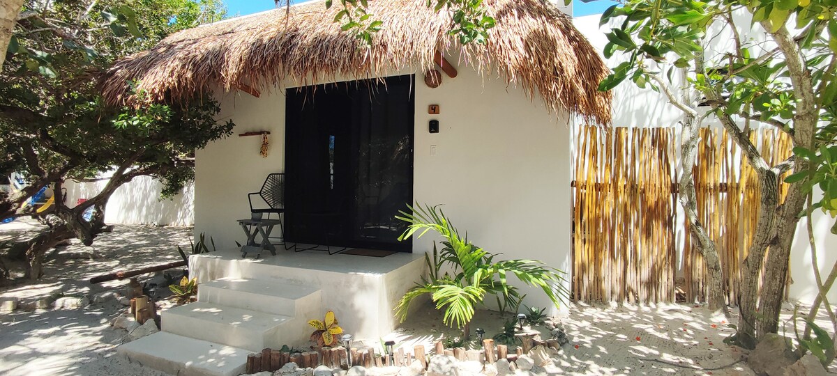 A charming casita sits beneath a thatched roof, featuring a spacious porch with two chairs. Lush greenery surrounds the entrance, framed by a wooden fence. Steps lead to a main door, providing access to a cozy and welcoming interior.