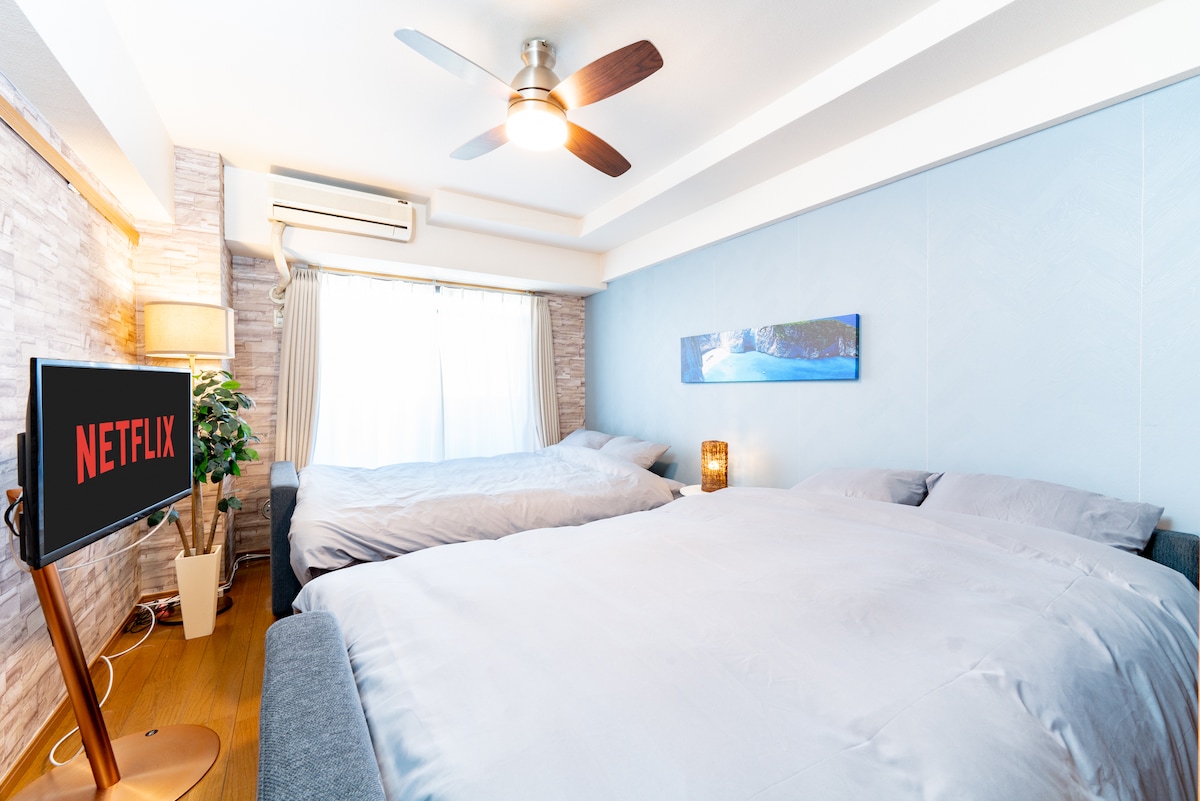 A twin bedroom is furnished with two spacious beds dressed in light grey bedding. A ceiling fan is centered above, and natural light filters through a large window with sheer curtains. A TV display nearby indicates access to streaming services.