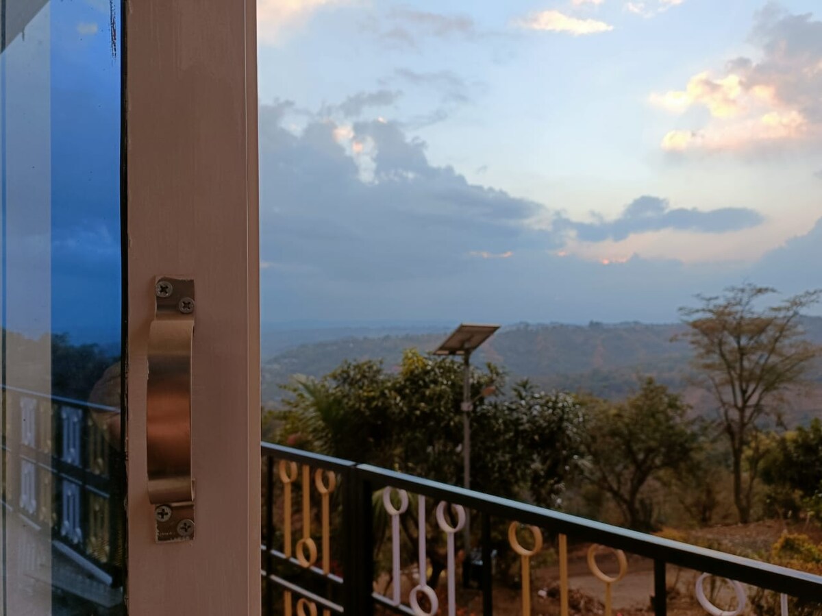A view from a doorway showcases a vast landscape with rolling hills under a cloudy sky. A balcony railing is visible in the foreground, while greenery and a solar panel are seen in the distance, contributing to the natural setting.