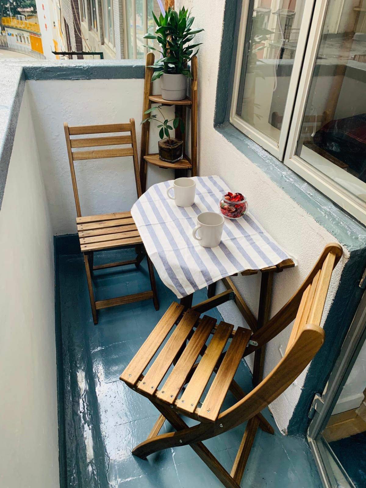 A compact balcony is presented with two wooden folding chairs and a small table covered by a striped cloth. A potted plant is positioned on a wooden shelf nearby, while two mugs await on the table, creating an inviting space for relaxation.