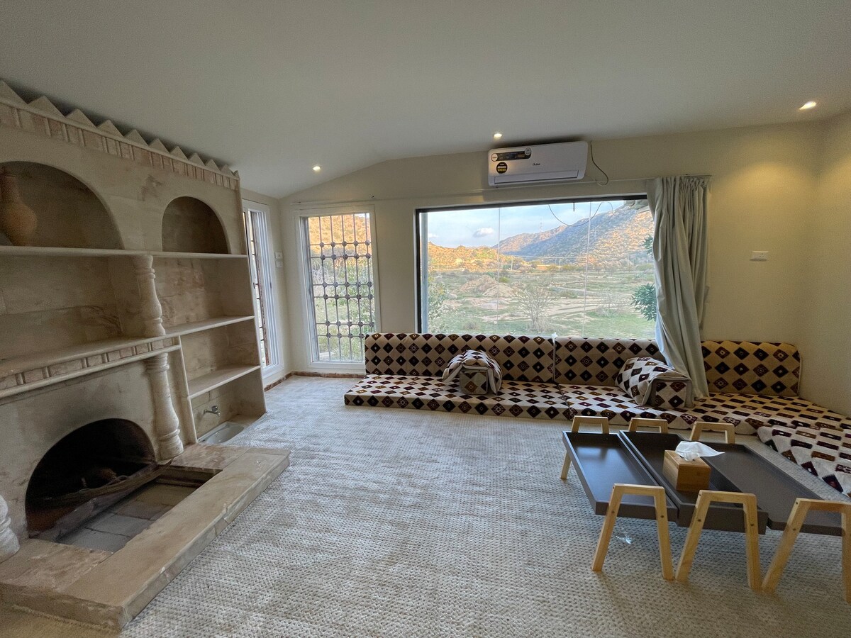 A cozy living area features a unique built-in fireplace and a wall of large windows providing scenic views of the surrounding mountains. A patterned seating area is accompanied by a low coffee table, creating a comfortable space for relaxation.