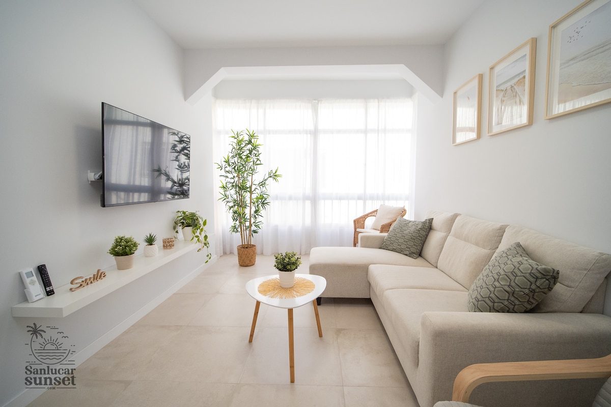 A light-filled living area features a comfortable sectional sofa adorned with decorative pillows. A round coffee table sits in front, accompanied by potted plants. A wall-mounted TV is positioned opposite the seating. Large windows covered by sheer curtains enhance the space's brightness.