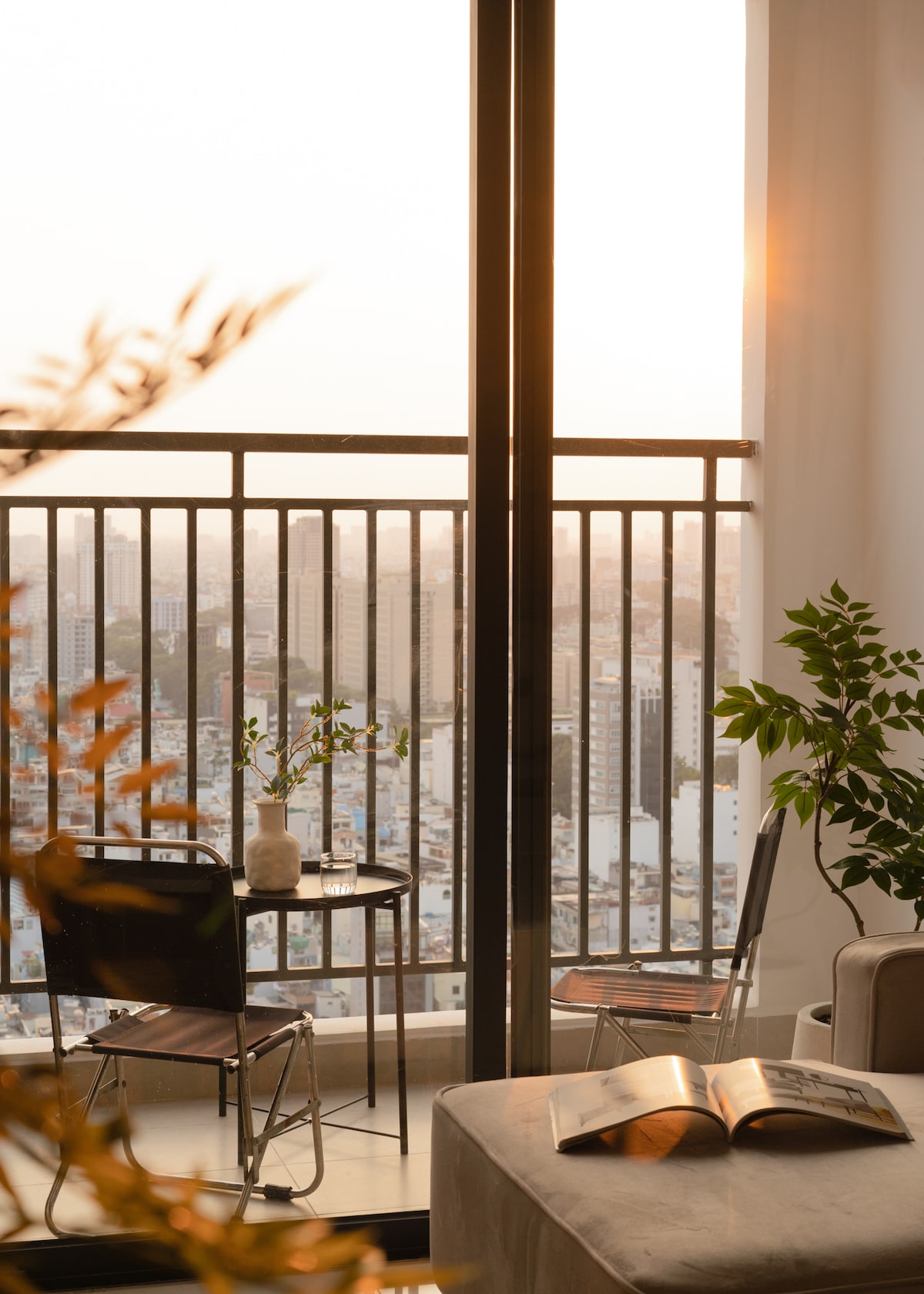 A spacious balcony is shown with a view of the city skyline at sunset. A small table holds a vase with greenery, and two modern chairs are positioned to enjoy the scenery. Natural light softly illuminates the interior, creating a serene atmosphere.