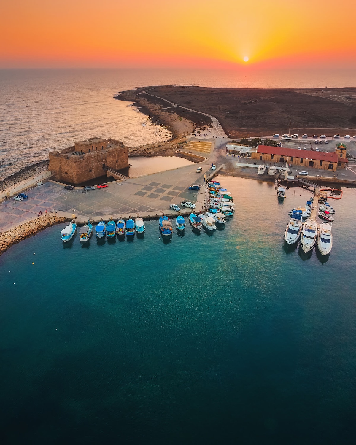 A serene coastal view captures a marina filled with boats, surrounded by calm waters. The sun sets on the horizon, casting warm hues across the sky. A historic structure stands near the shore, with nearby buildings lining the coastline.