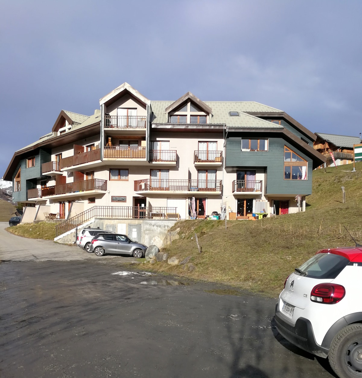A three-story building with a sloped roof features multiple balconies and large windows, showcasing an appealing architectural design. The surrounding area is grassy, and several parked cars are visible in the foreground, indicating available private parking for guests.