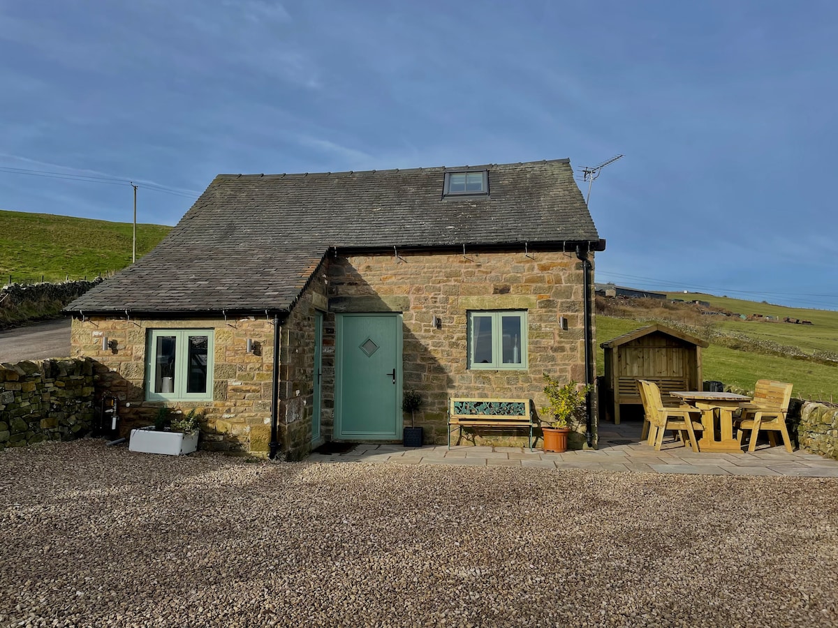A stone cottage is showcased against a clear blue sky, featuring a slate roof and a light green door. Windows are visible on both levels, while a patio area with outdoor seating is located to the right. The surrounding landscape consists of gentle hills.