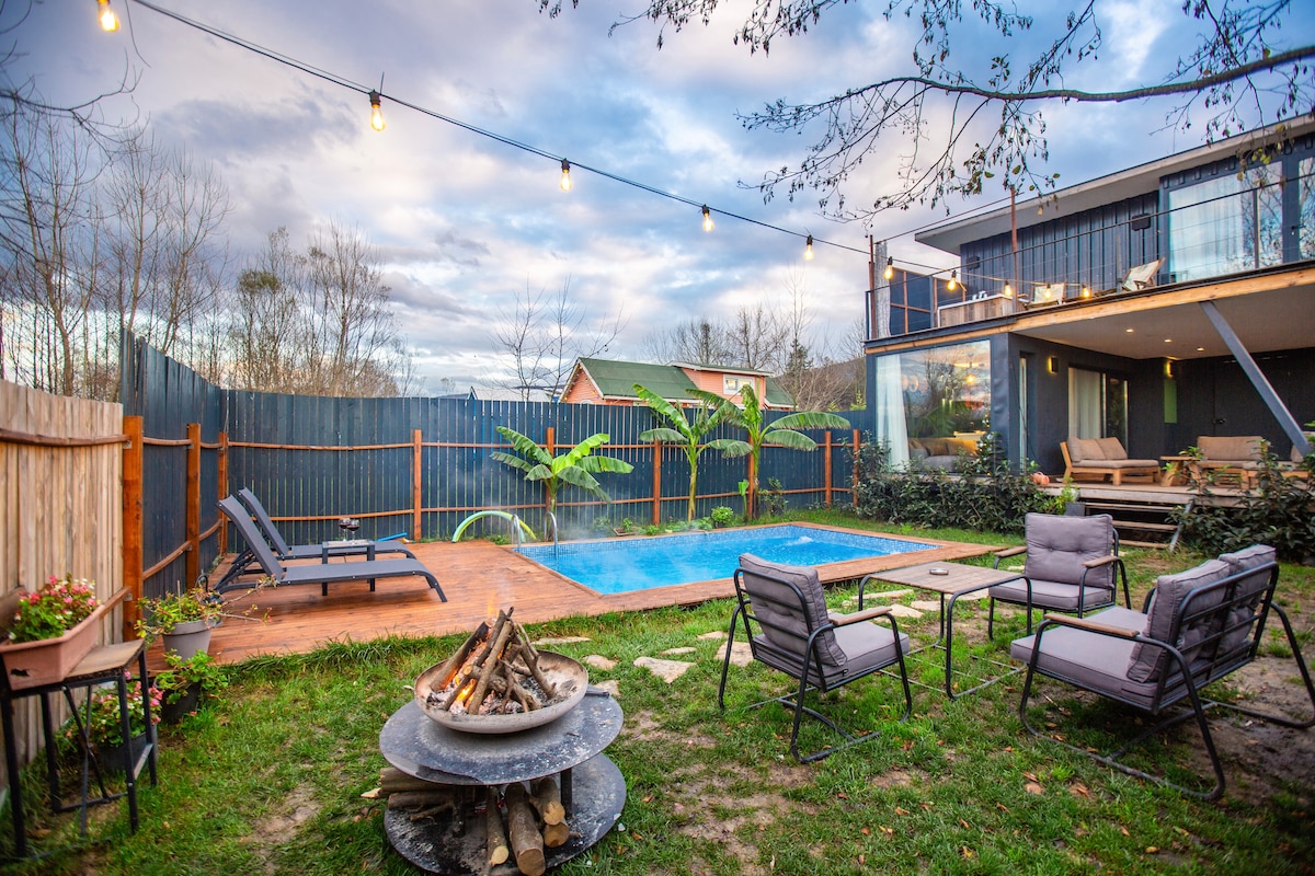 An outdoor area features a small swimming pool surrounded by lounge chairs, with a wooden deck and a fire pit at the center. Foliage lines the perimeter, and overhead, string lights are hung, creating a relaxed atmosphere.