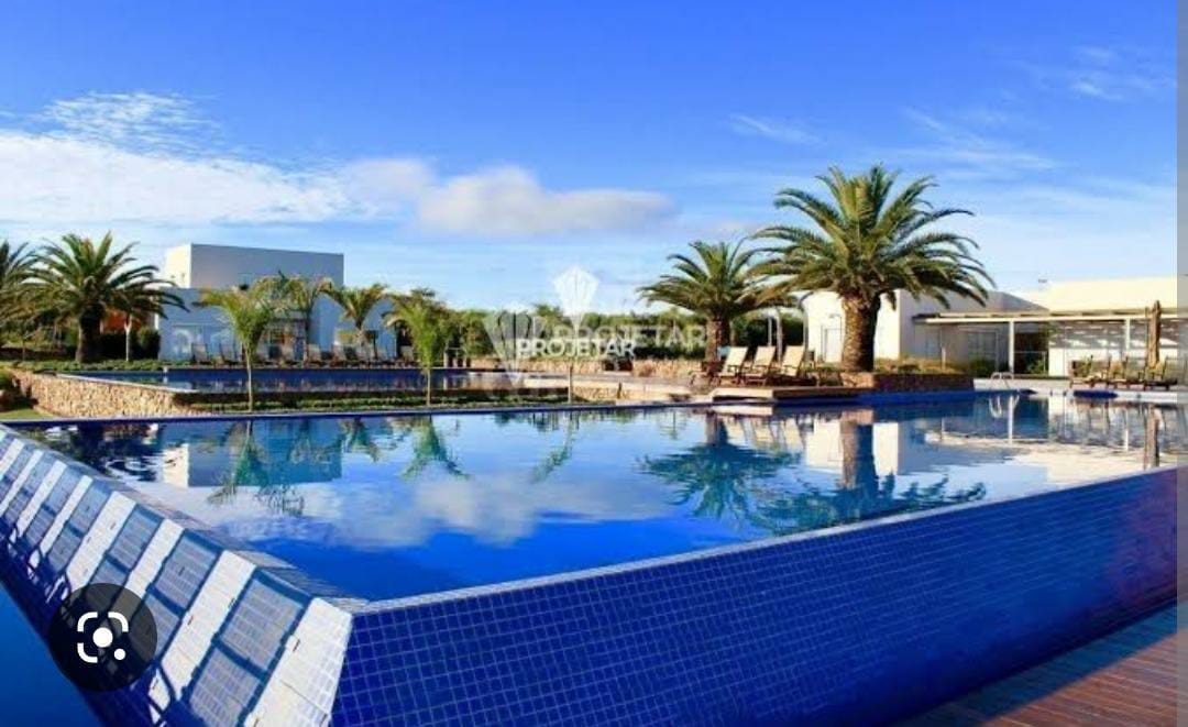 A large outdoor swimming pool is surrounded by tropical palm trees, reflecting a clear blue sky. Lounge chairs are positioned around the pool area, creating spaces for relaxation. The serene landscape is complemented by the architecture of nearby buildings.