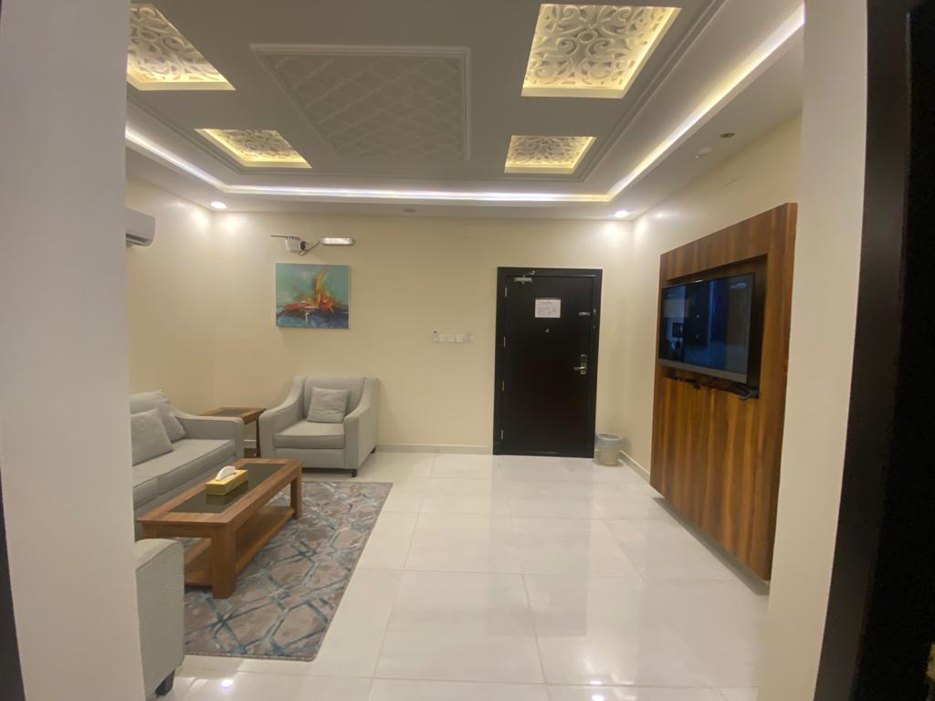 A modern living area features light-colored sofas arranged around a wooden coffee table. A large flat-screen TV is mounted on an accent wall, complemented by a subtle piece of artwork. Soft lighting is provided by elegantly designed ceiling fixtures, reflecting off the glossy floor.