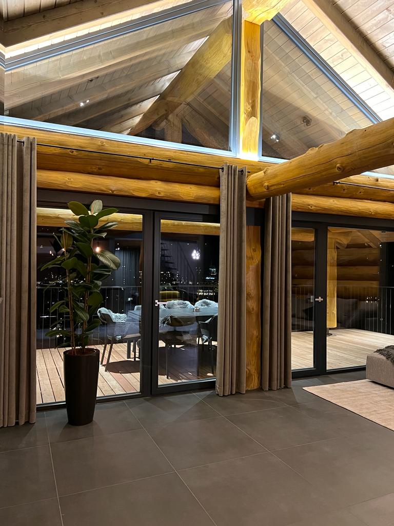 The spacious living area is highlighted by a double high ceiling with exposed wooden beams. Large glass doors lead to an outdoor terrace furnished with a dining table. Natural light floods the room, complemented by decorative plants and soft drapes on the windows.
