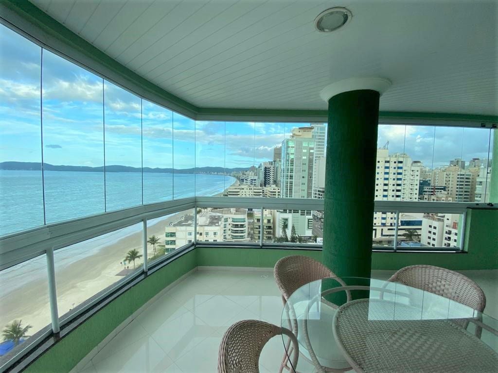 A spacious balcony features glass railing, offering unobstructed views of the coastline and cityscape. A round glass table is accompanied by four wicker chairs, providing a comfortable space to enjoy the scenery. The ocean glimmers in the distance under a partly cloudy sky.