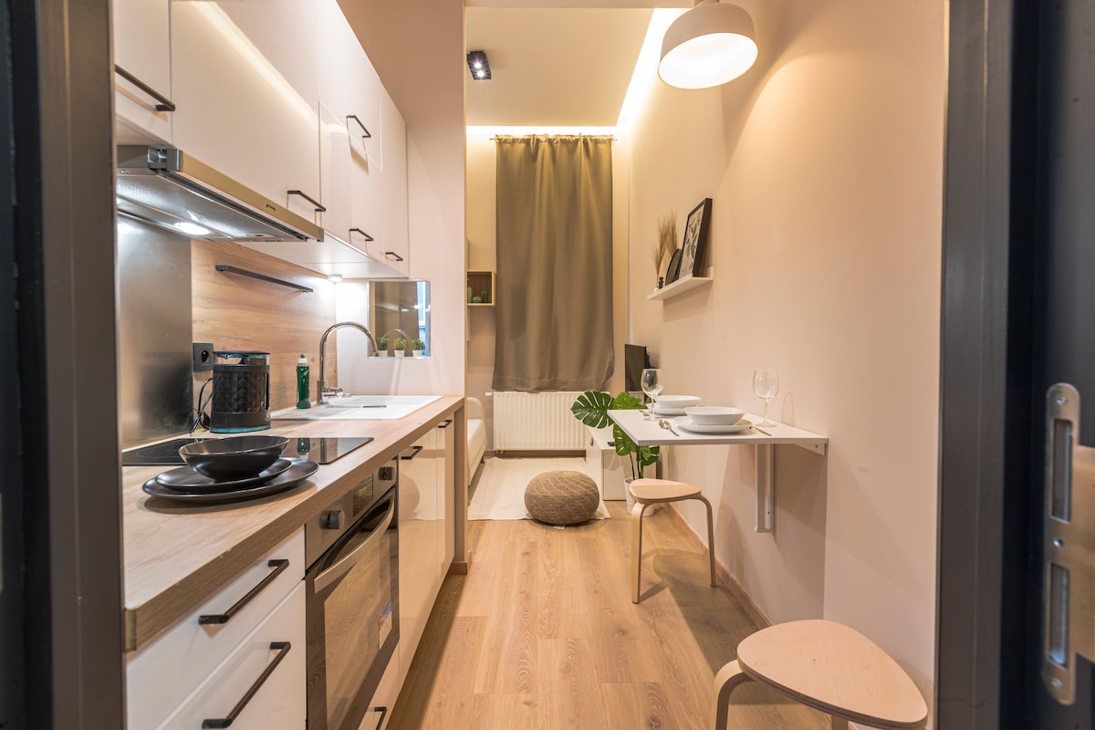 A compact kitchen space features modern cabinetry and integrated appliances. A small dining area with a wall-mounted table and two chairs is present. Natural wood tones and soft lighting contribute to a warm ambiance. A potted plant adds a touch of greenery near the floor.