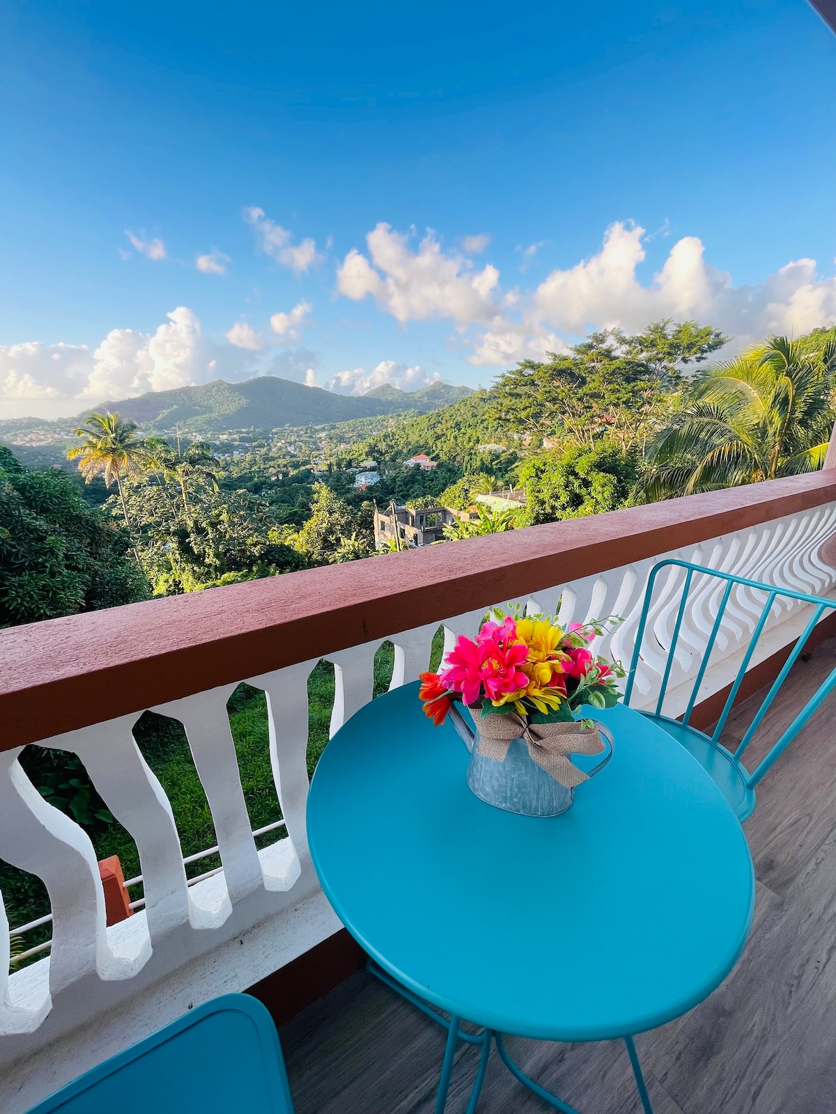 A balcony is presented with a vibrant blue table and matching chairs, adorned with a vase of fresh flowers. Lush greenery and mountains fill the background under a partially cloudy sky, offering a serene view of the natural surroundings.