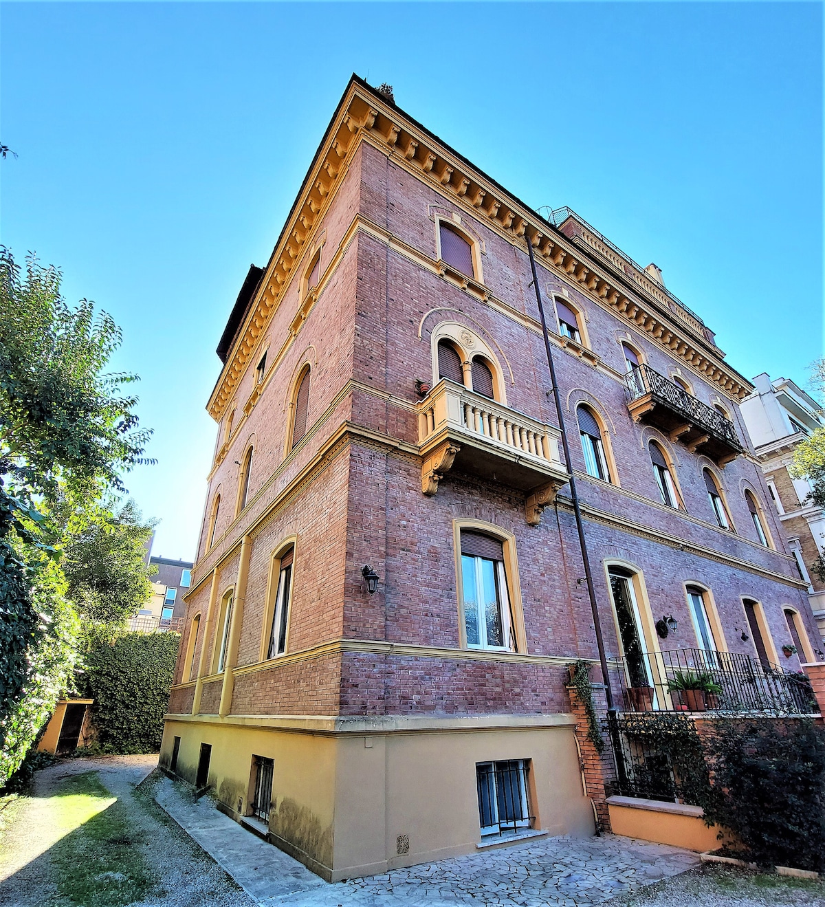 A charming brick villa from the early 1900s is presented, featuring multiple balconies and large windows. The structure is framed by greenery, with a stone pathway leading to the entrance, providing a blend of historical architecture and inviting outdoor space.
