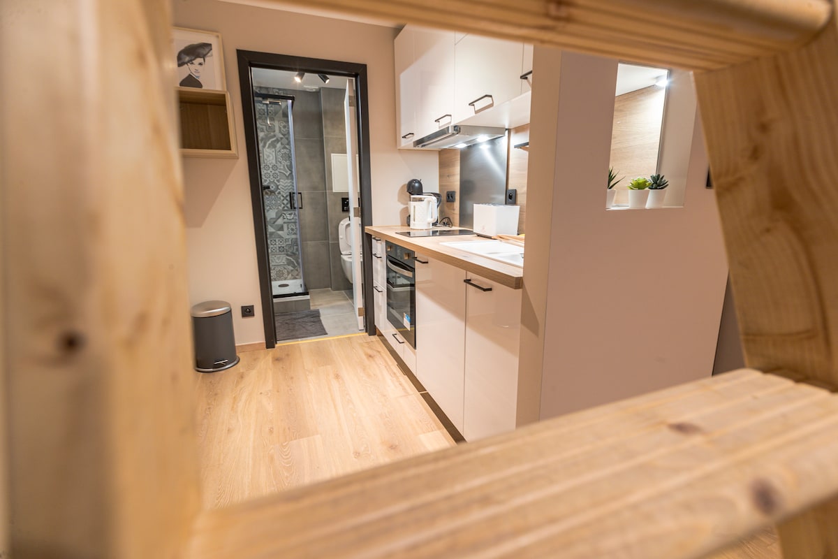 A well-equipped kitchen area is visible, featuring modern cabinetry and appliances. Light wood flooring complements the contemporary design. A doorway leads to a bathroom at the back, with elements of transparency and clean lines enhancing the spacious feel.