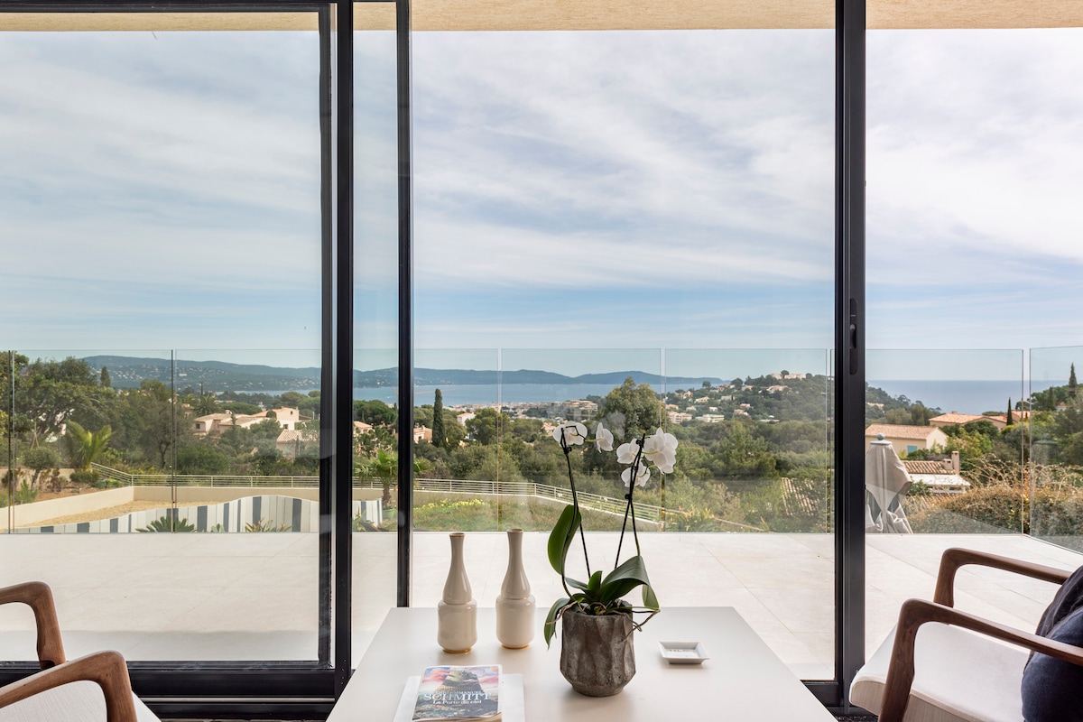 A modern living space features large glass doors, providing a panoramic view of the landscape and sea. A white table is set with a plant and two vases, while a magazine rests on the table. Comfortable seating is visible in the foreground.