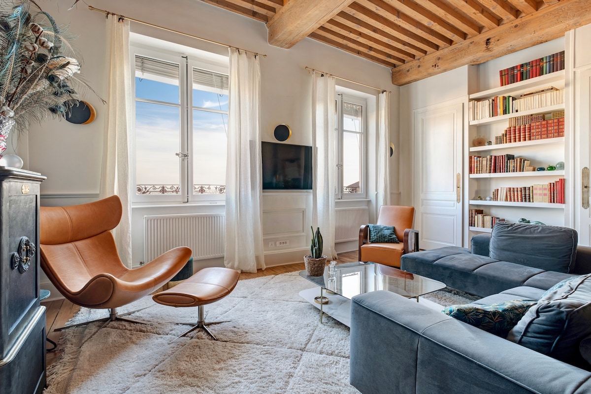 A living area is furnished with a sleek leather lounge chair and a modern sectional sofa. A glass coffee table is situated in the center, surrounded by bookshelves lined with various titles. Natural light fills the space through large windows dressed in sheer curtains.