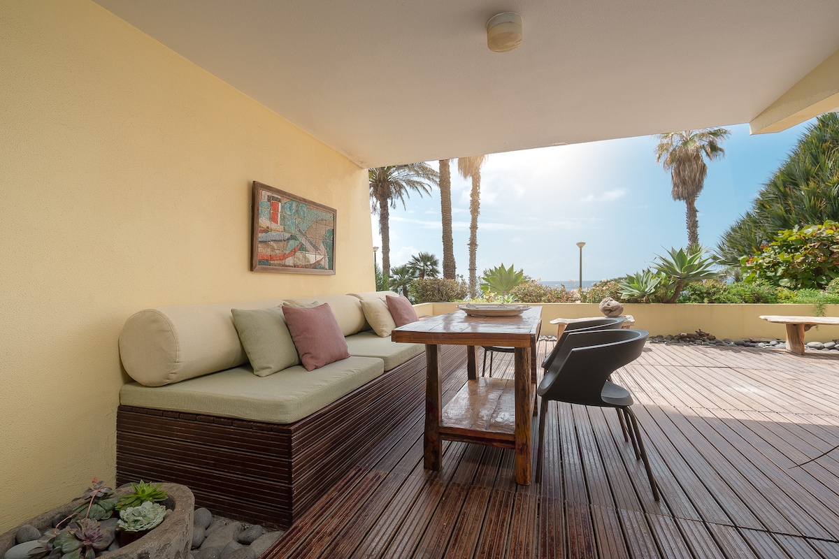 A spacious patio area features a wooden dining table accompanied by modern chairs. A comfortable seating arrangement with plush cushions is visible, set against a backdrop of greenery and palm trees. Natural light filters in, enhancing the relaxed outdoor ambiance.