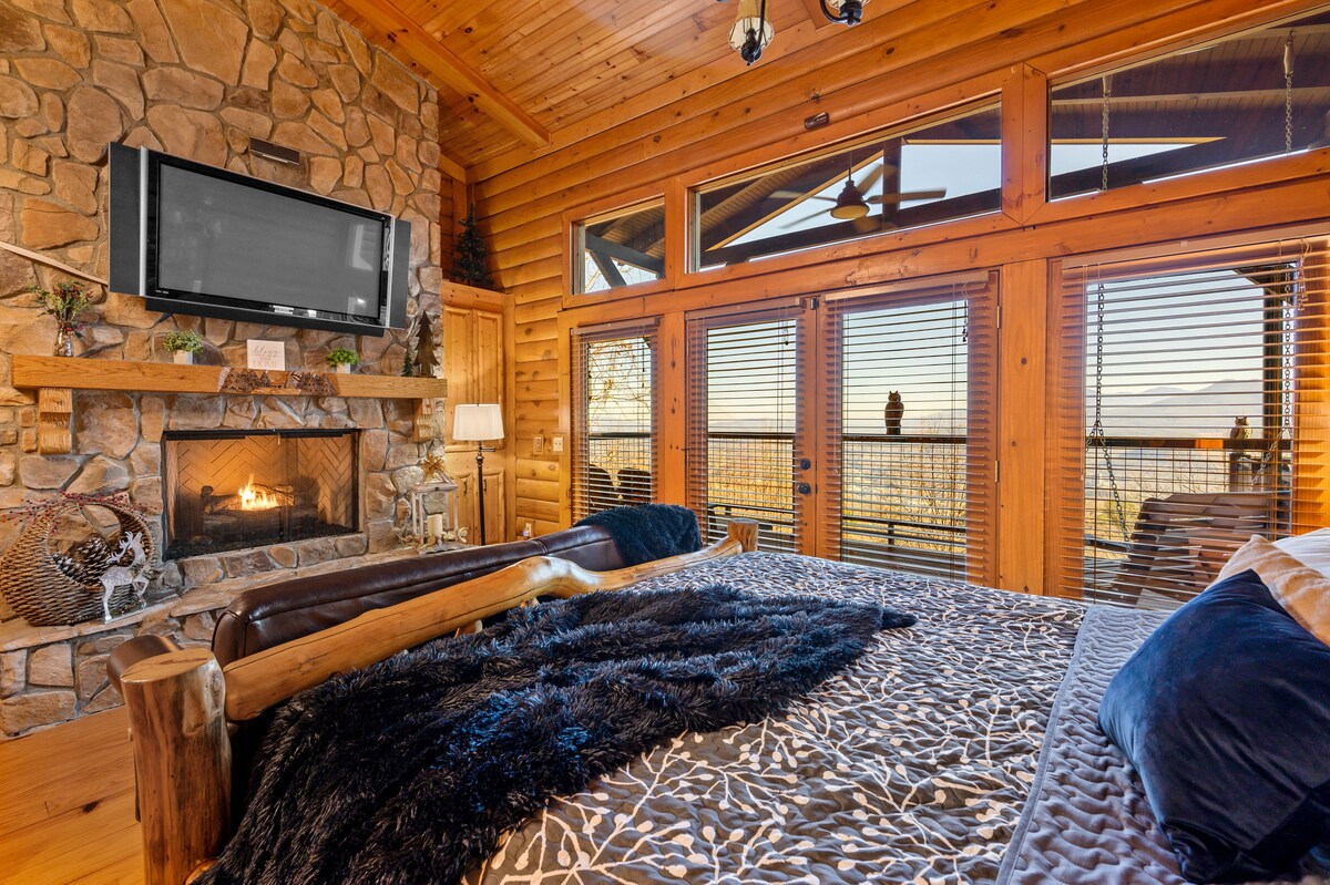 A warm bedroom features a spacious bed adorned with a textured blanket. A stone fireplace is positioned beneath a television, and expansive windows offer views of the outside deck and Smoky Mountains. Natural wood accents enhance the inviting atmosphere.