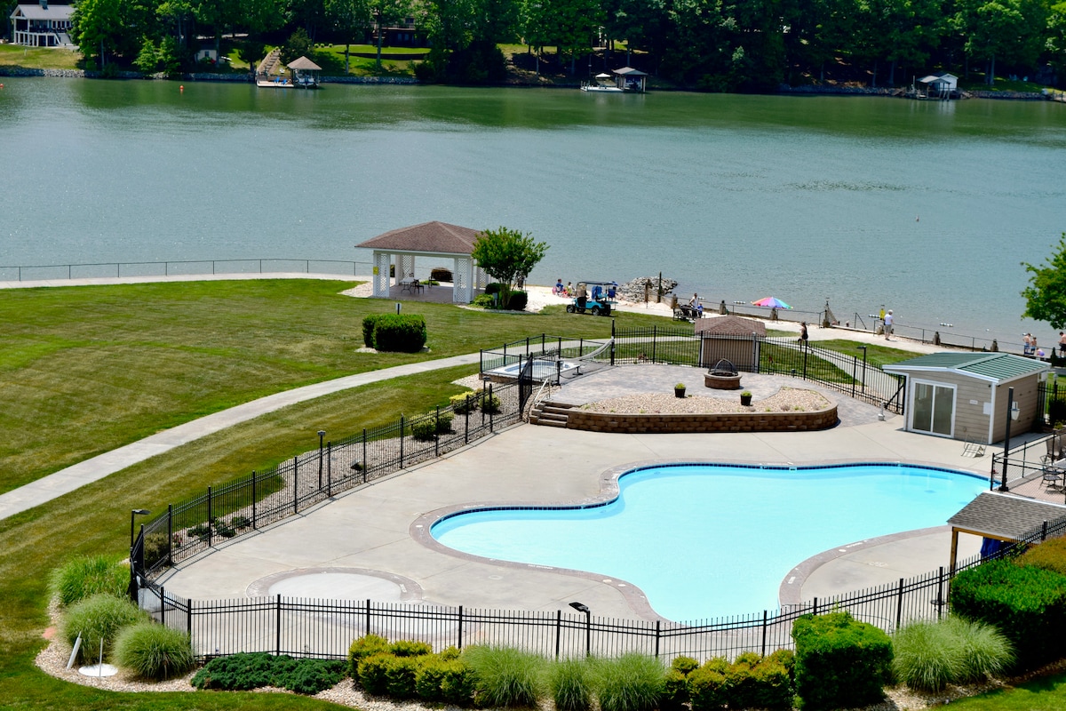 A clear view of a large outdoor pool surrounded by a gated patio area. Lush green grass and shrubs line the perimeter. In the background, a serene lake is visible, along with a gazebo and people enjoying the shoreline.