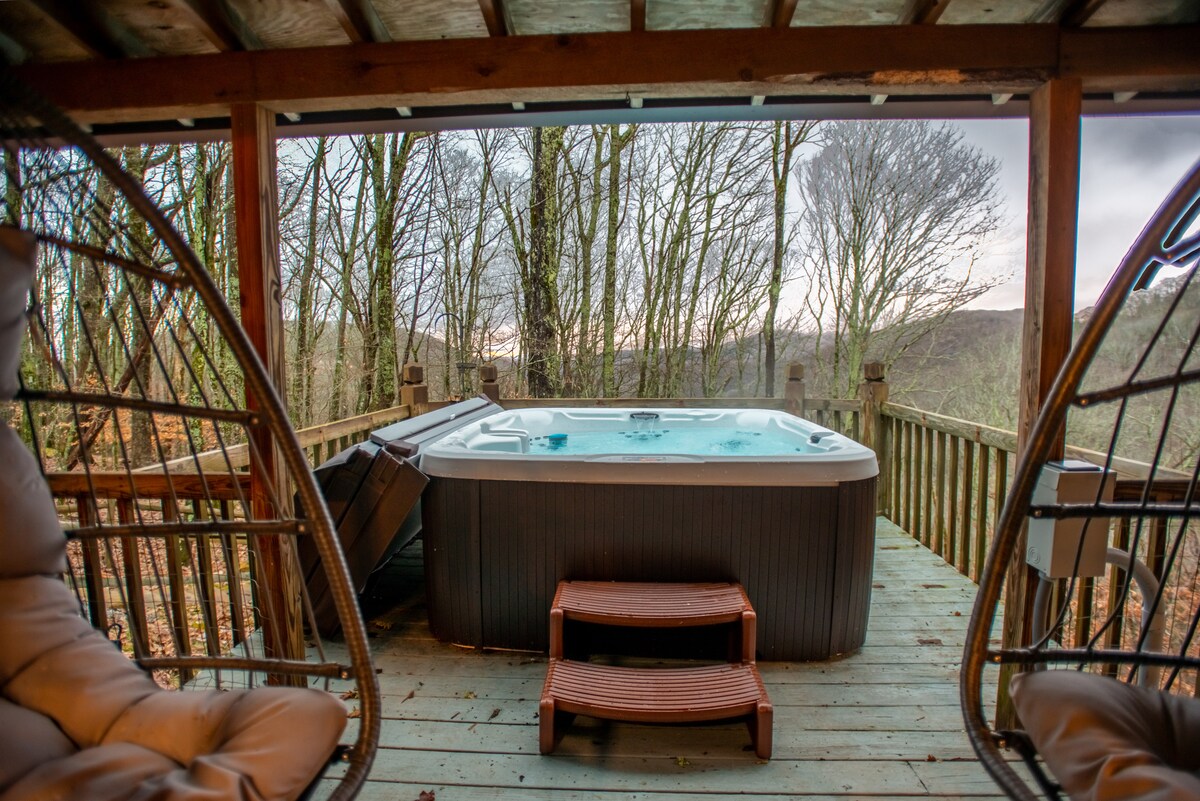 A hot tub is positioned on a wooden deck, surrounded by trees and offering serene views of the mountain landscape. Two hanging chairs are visible nearby, providing a comfortable seating area to enjoy the tranquil surroundings.