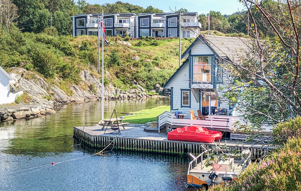 A charming house sits by the calm water, featuring a dock with seating and a red inflatable kayak. The backdrop includes several modern buildings, and greenery surrounds the area, offering a tranquil setting for outdoor activities.