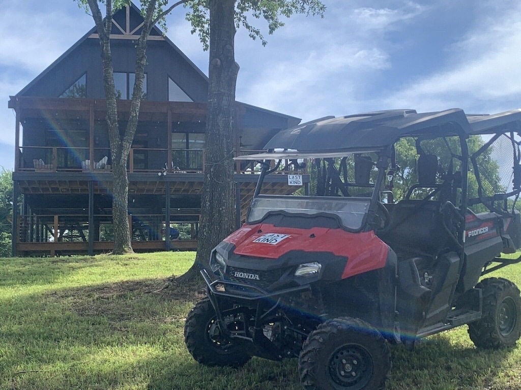 An all-terrain vehicle is parked on a grassy area, featuring a bright red exterior. In the background, a modern cabin is visible, elevated on stilts and surrounded by trees. The cabin's large windows provide a view of the interior spaces.