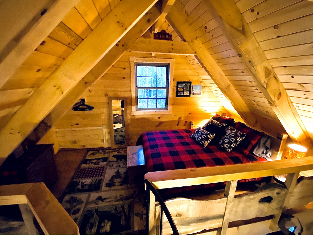 An open loft area features a spacious bed adorned with a plaid blanket, surrounded by natural wood paneling. Decorative items and a framed photo add character, while a small window allows for natural light. A cozy ambiance is created by soft lighting throughout the space.
