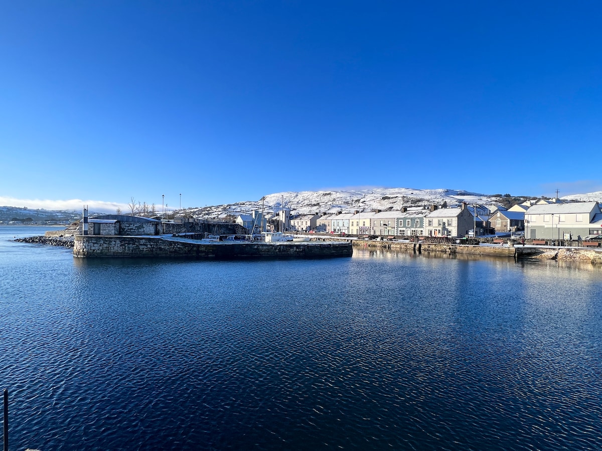 An expansive view of a calm harbor reflects the azure sky. Quaint buildings line the waterfront, with a backdrop of gently rolling hills. The image captures the serene environment, showcasing boats docked along the pier, inviting a sense of tranquility and exploration.