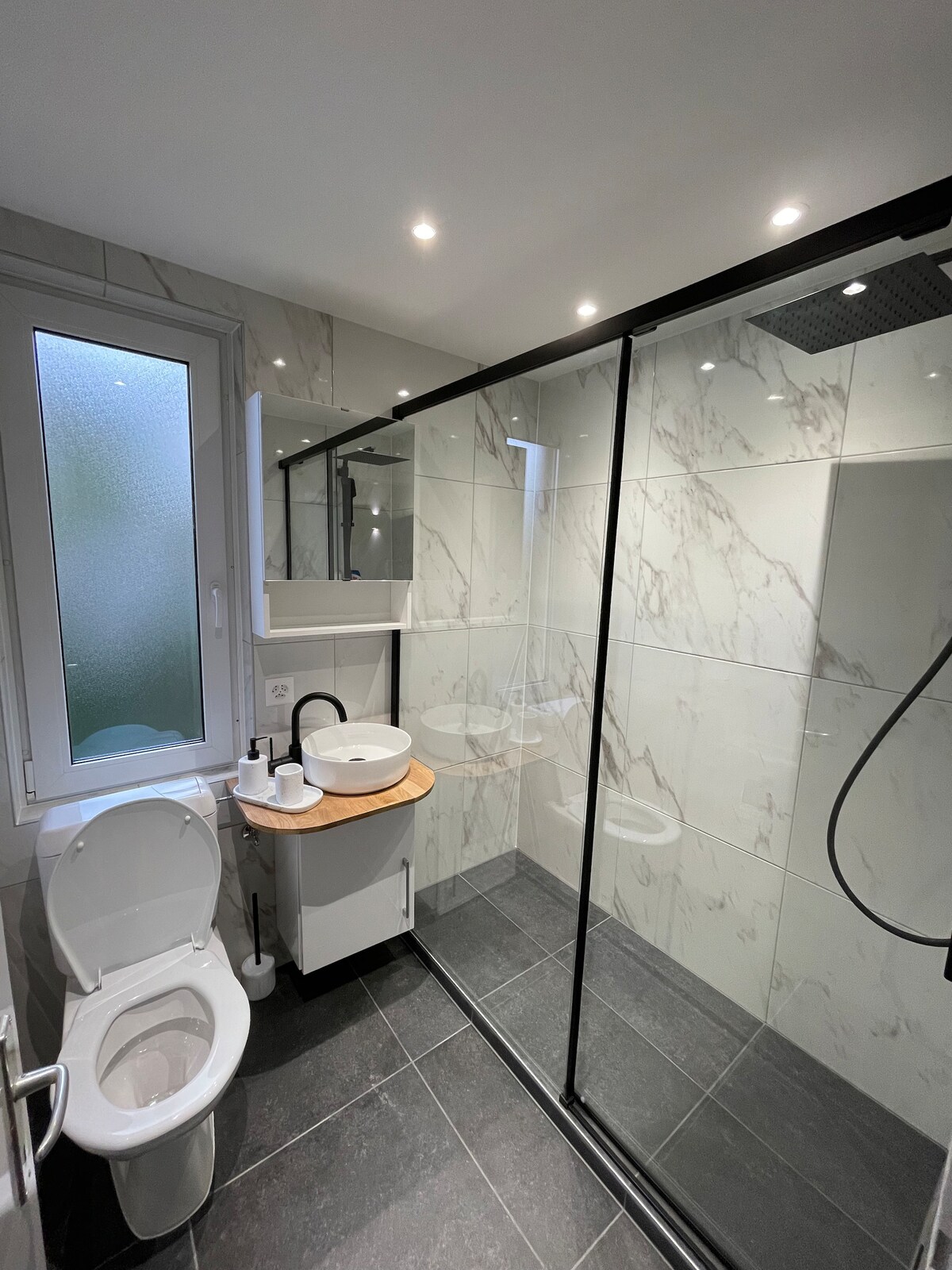 A modern bathroom is shown with a spacious walk-in shower enclosed by glass. A circular sink is mounted on a wooden countertop. Natural light comes through a window with frosted glass, while grey tiles create a clean, minimalist look.