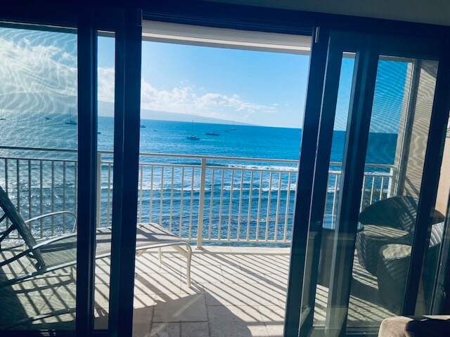 An open balcony view showcases the ocean and horizon, with gentle waves visible. The glass doors lead to the outdoors, where patio furniture offers a place to relax. Sunlight creates a shimmering effect on the water, reflecting the clear blue sky.