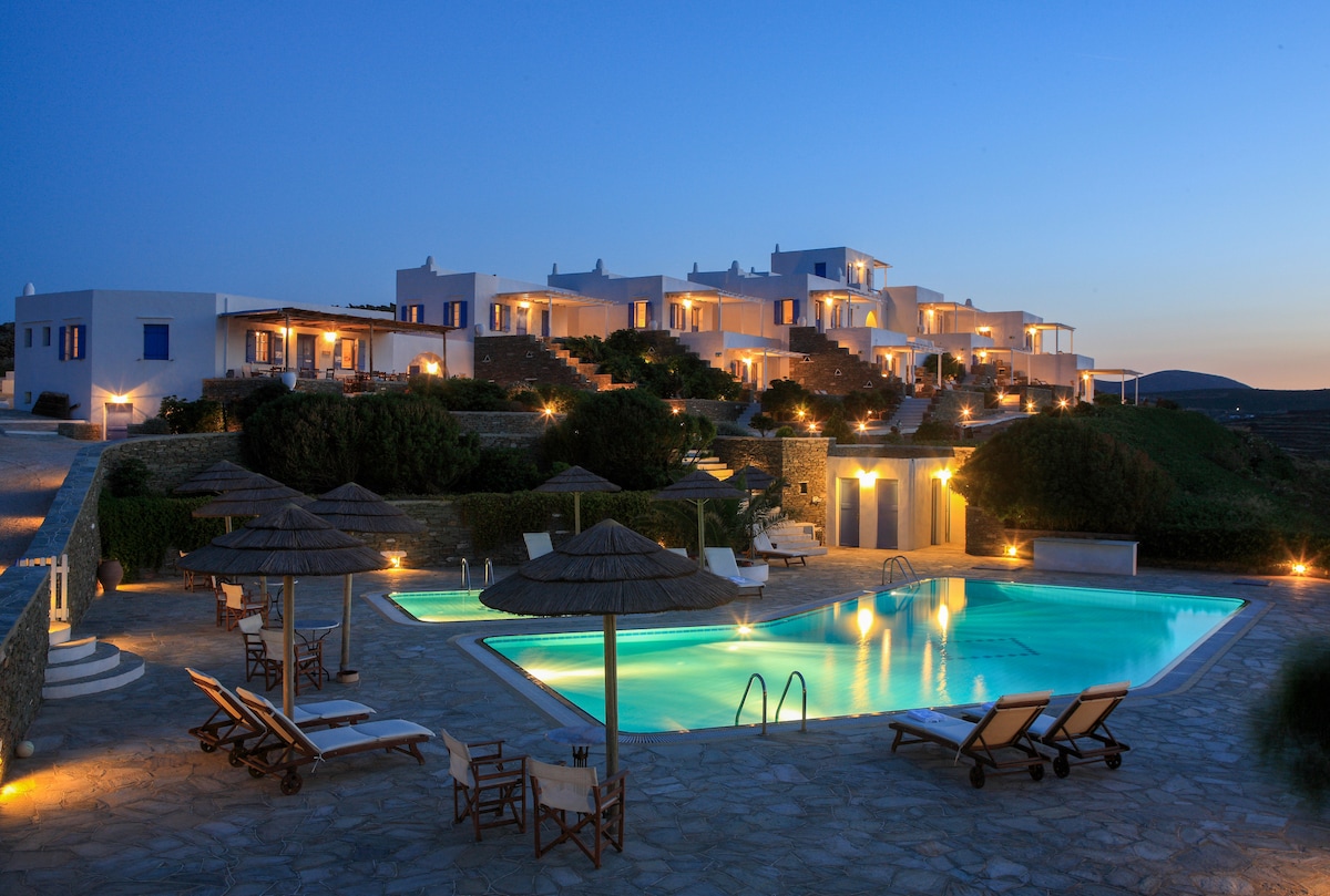 A serene evening scene showcases a modern Cycladic maisonette illuminated by soft lighting. An inviting swimming pool reflects the surroundings, surrounded by lounge chairs and shaded umbrellas. The landscape features well-kept greenery leading up to the two-level structure that houses multiple independent units.