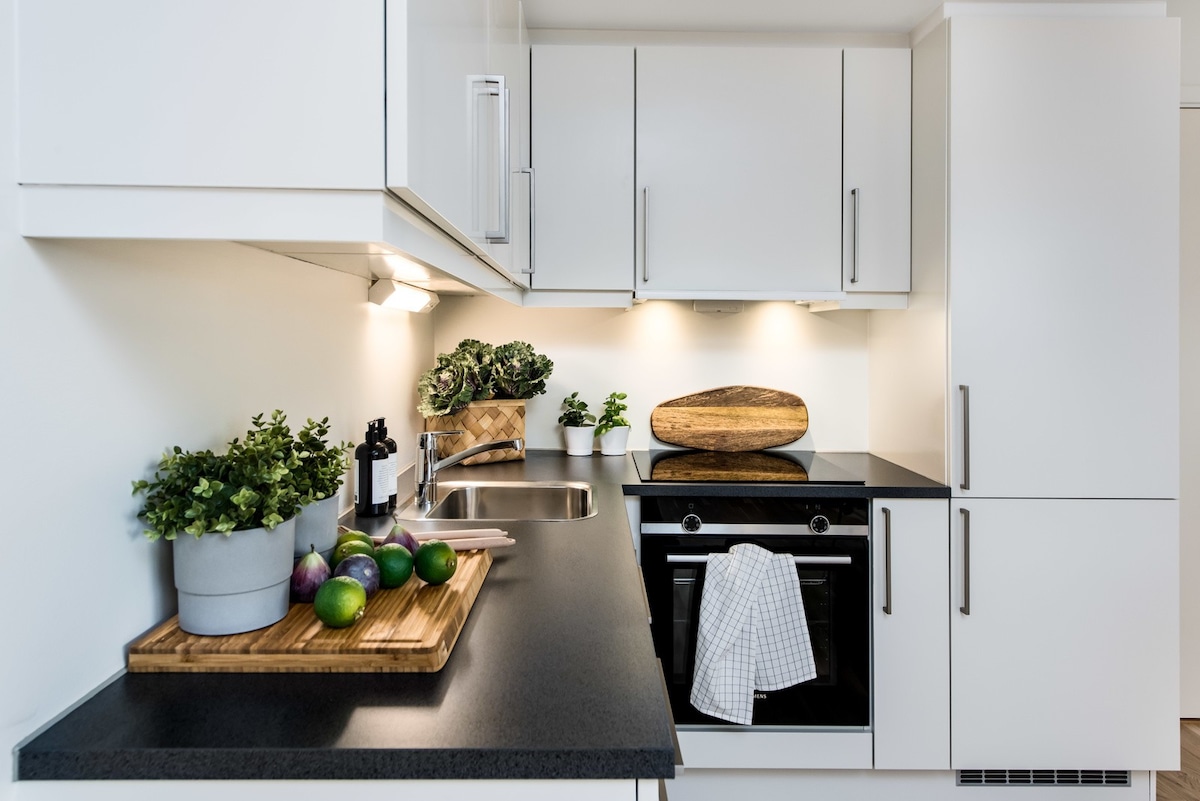 A modern kitchen features sleek white cabinetry and a dark countertop. Potted herbs and a wooden cutting board with fruits are arranged on one side. An oven and stove are positioned beneath warm lighting, providing a well-lit preparation area.
