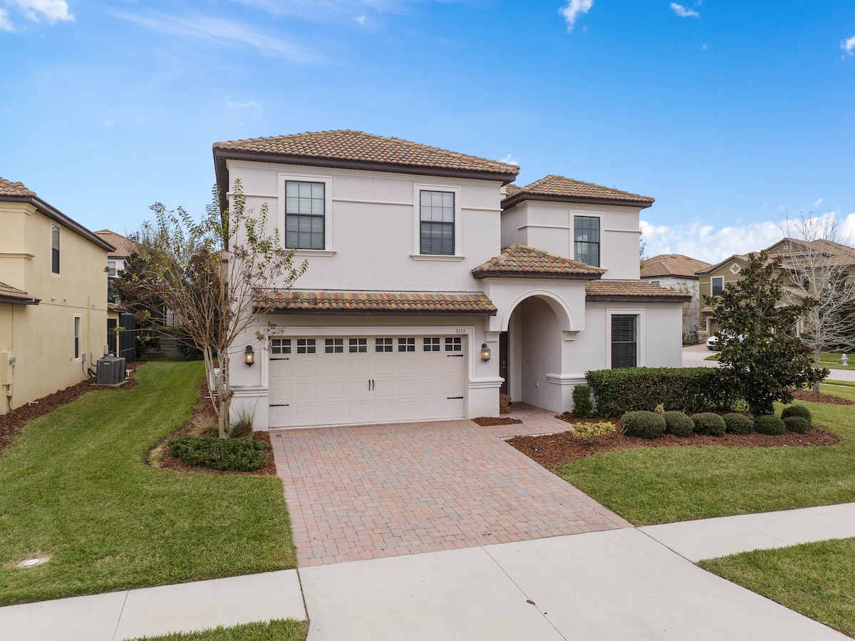A modern two-story home is presented, featuring a tile roof and a spacious paver driveway. Lush greenery lines the front yard, complementing neat shrubs. Windows adorned with shutters enhance the facade, while a covered entrance invites guests into the residence.